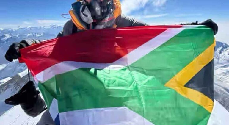 A person wearing cold-weather gear and an oxygen mask holds the South African flag at a snowy mountain summit with a clear sky and distant peaks in the background.