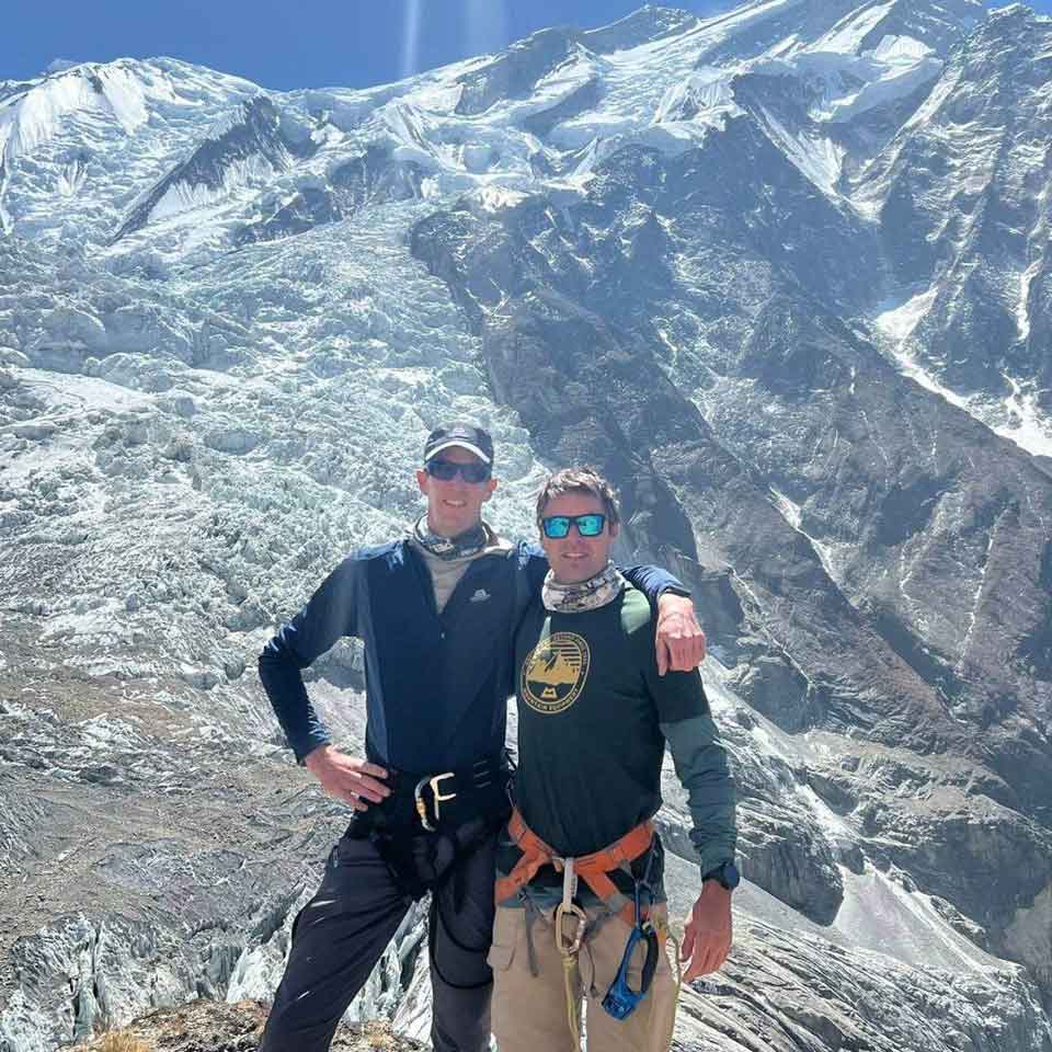Two climbers wearing sunglasses and mountaineering gear stand arm-in-arm on a rocky, snowy mountainside with a towering, sunlit glacier and blue sky in the background.