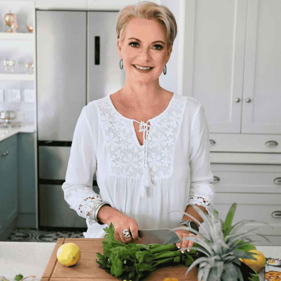 A smiling woman with short blonde hair stands in a bright kitchen, wearing a white blouse and chopping celery on a wooden cutting board with a lemon and pineapple nearby.