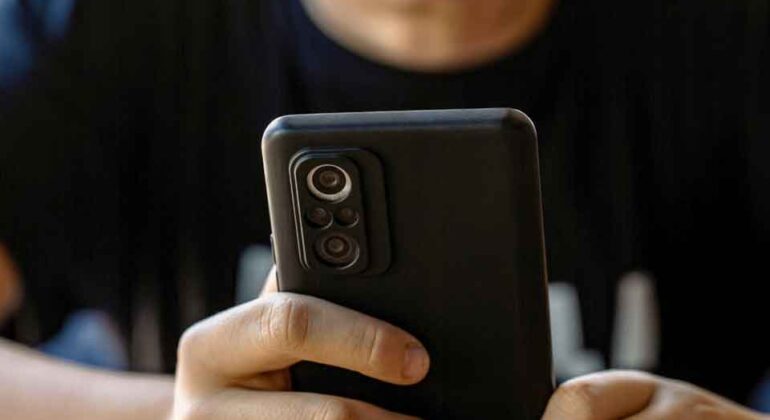 A child sits at a table holding a smartphone with both hands, focusing intently on the screen. The background is blurred.