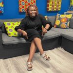 A woman with curly hair sits on a grey couch, smiling and pointing at her decorated sandals. The couch has colorful patterned pillows and vibrant fabric art on the blue wall behind her.