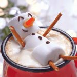 A mug of hot chocolate topped with foam, decorated with a snowman made of three marshmallows, pretzel stick arms and legs, a candy corn nose, and chocolate details for eyes and a smile.