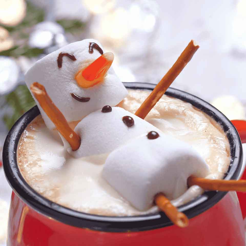 A mug of hot chocolate topped with foam, decorated with a snowman made of three marshmallows, pretzel stick arms and legs, a candy corn nose, and chocolate details for eyes and a smile.