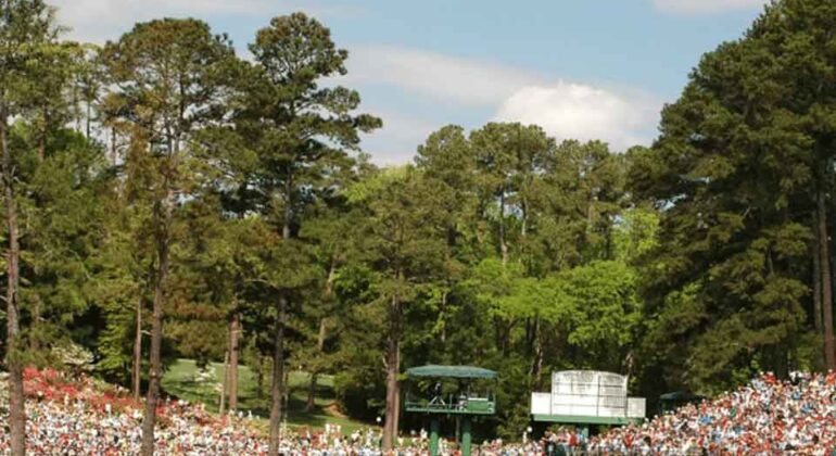 A large crowd watches a golf tournament on a green course surrounded by tall trees, under a partly cloudy sky. Several people stand on the putting green near the flagstick and a sand bunker.