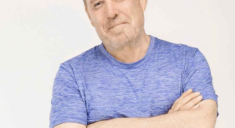 A middle-aged man with short hair wearing a blue t-shirt stands with his arms crossed, smiling slightly, against a plain light background.