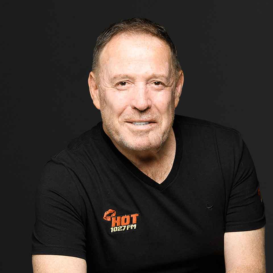 A smiling middle-aged man with short hair and a trimmed beard wears a black t-shirt with a HOT 102.7 FM logo, posing against a plain, dark background.