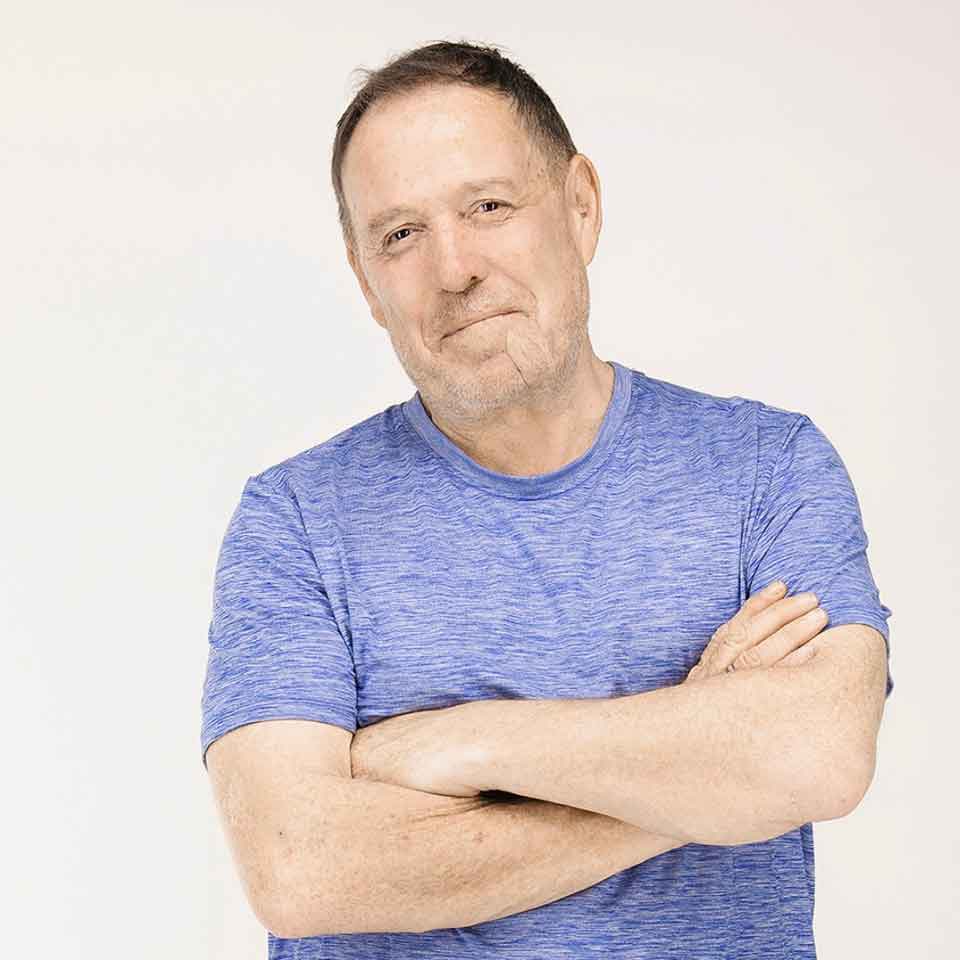 A middle-aged man with short hair wearing a blue t-shirt stands with his arms crossed, smiling slightly, against a plain light background.