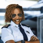 A smiling woman wearing a pilot uniform and aviator sunglasses stands confidently with arms crossed in front of an airplane.