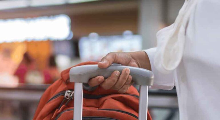 A person wearing a white shirt holds the handle of an orange suitcase at an airport, with blurred people and lights in the background.