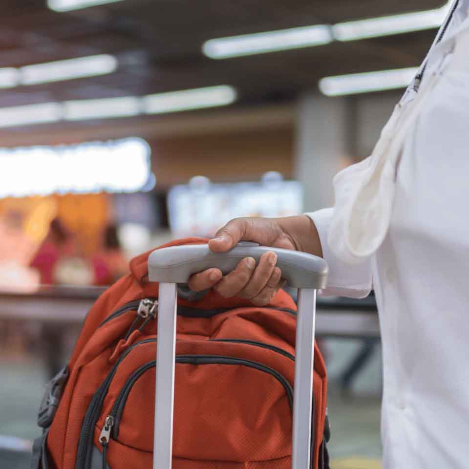 A person wearing a white shirt holds the handle of an orange suitcase at an airport, with blurred people and lights in the background.