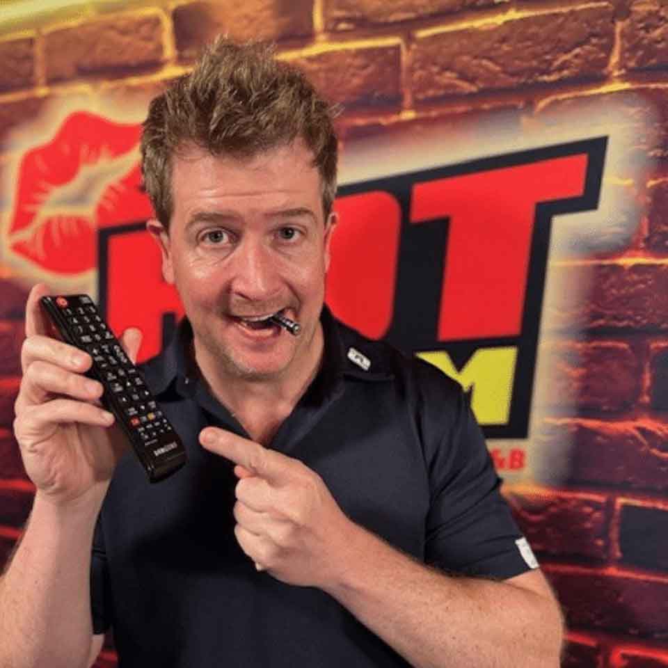 A man in a black shirt smiles while holding a TV remote and pointing at it, with a pen in his mouth. He stands in front of a brick wall with a large HOT FM logo in red and yellow.