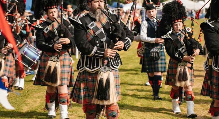A group of people in traditional Scottish attire, including kilts and feathered hats, play bagpipes and drums while marching outdoors on a grassy field during a cultural event or festival.