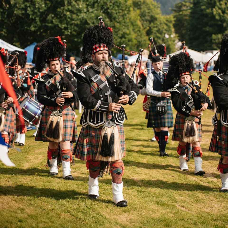 A group of people in traditional Scottish attire, including kilts and feathered hats, play bagpipes and drums while marching outdoors on a grassy field during a cultural event or festival.