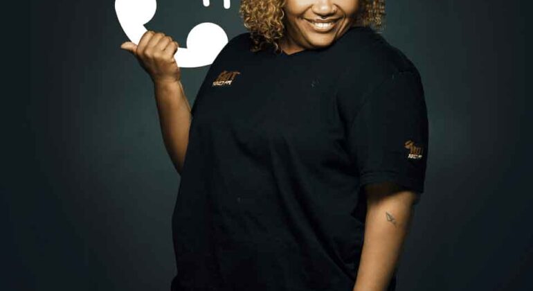 A smiling woman with curly hair wearing a black outfit poses against a dark background, holding up her hand in a phone gesture next to a white phone icon illustration.