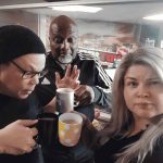 Three adults stand indoors holding mugs, posing for a selfie. One man wears glasses and a beanie, another man in a tracksuit gestures with his hand, and a woman with long, light hair looks at the camera.