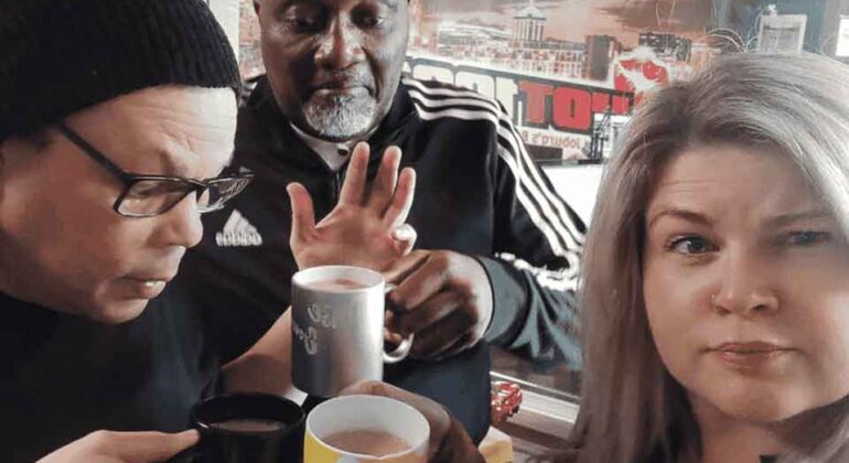 Three adults stand indoors holding mugs, posing for a selfie. One man wears glasses and a beanie, another man in a tracksuit gestures with his hand, and a woman with long, light hair looks at the camera.