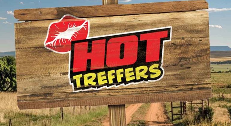 A wooden sign with a red lipstick mark and bold text reading HOT TREFFERS stands by a rural dirt road, with fields and blue sky in the background.