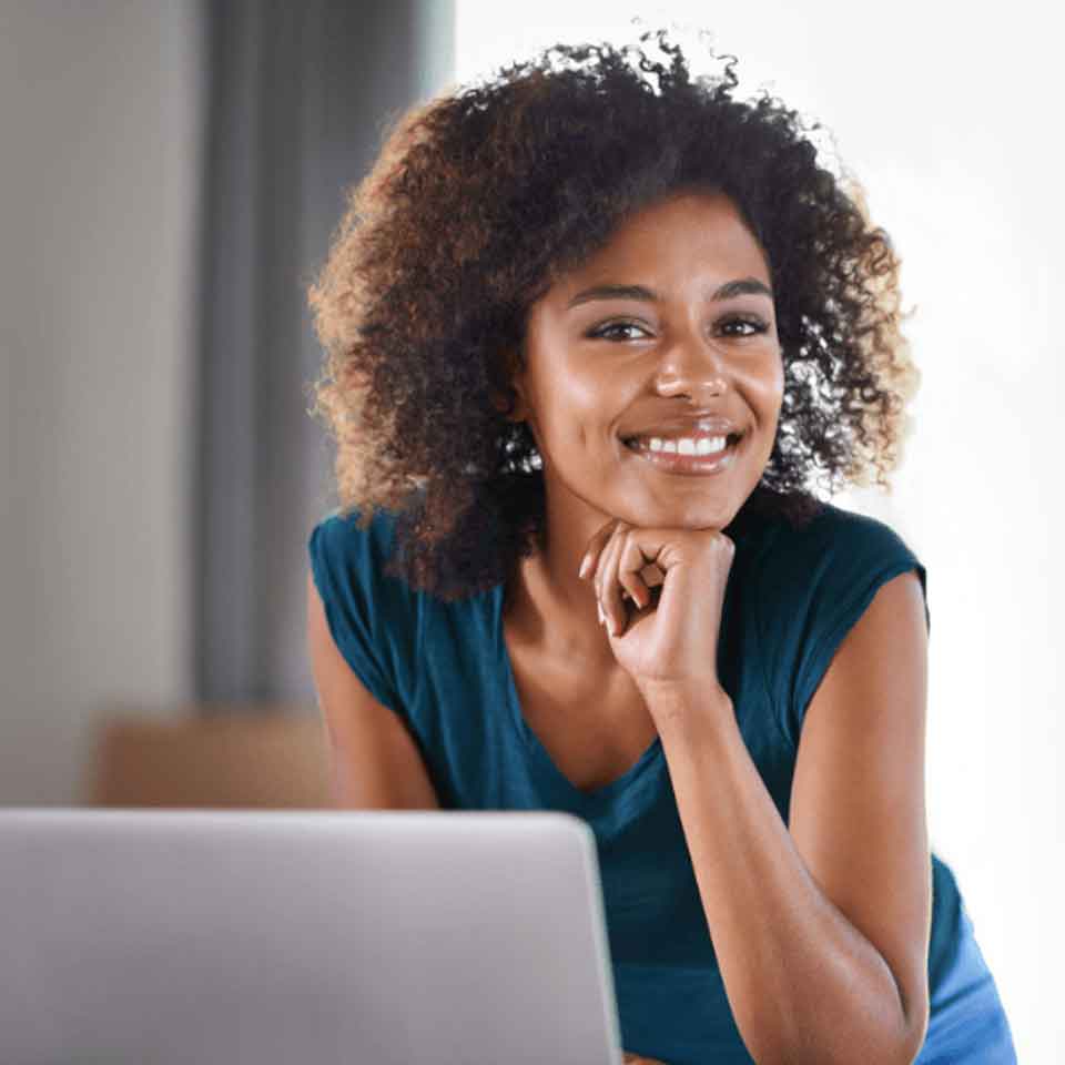 A woman with curly hair wearing a blue shirt smiles while sitting at a desk with a laptop, resting her chin on her hand.
