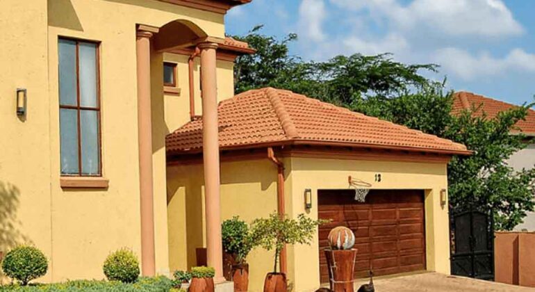 A modern house with a yellow exterior, red-tiled roof, large windows, and a well-maintained lawn with shrubs. A basketball hoop is mounted above the garage door, and the sky is blue with scattered clouds.