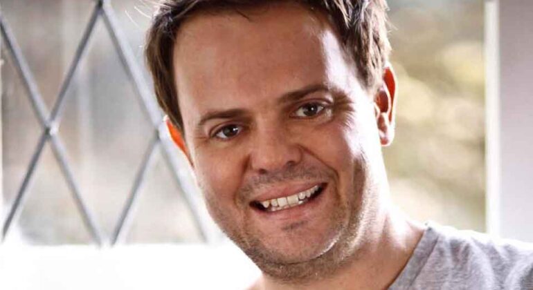 A man with short brown hair and a light beard smiles at the camera. He is wearing a light gray t-shirt and is sitting indoors near a window with a diamond-patterned frame.