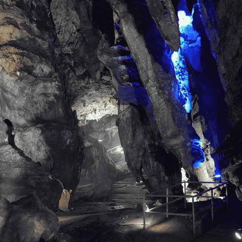 A rocky cave interior with jagged stone formations, illuminated by blue and white lights, evokes the mysterious beauty of the Sterkfontein Caves. A walkway with railings leads deeper into the cavern’s shadowy depths.
