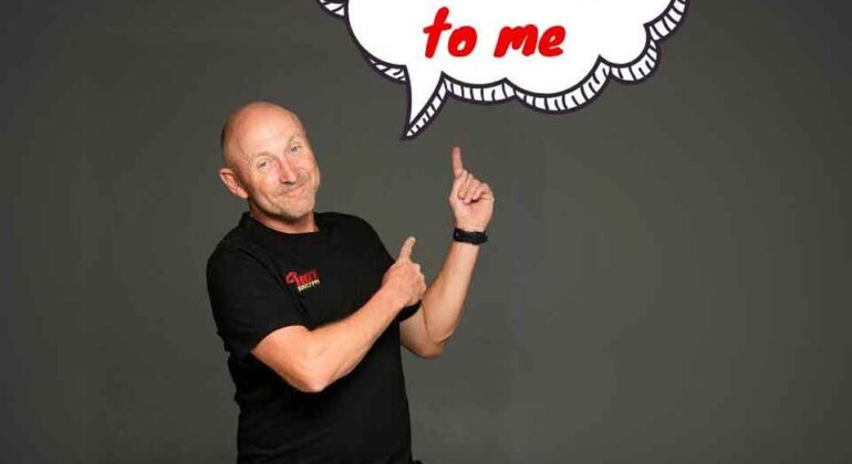 A bald man in a black t-shirt and jeans stands against a dark background, smiling and pointing up at a cartoon speech bubble that says, Talk dirty to me in red letters.
