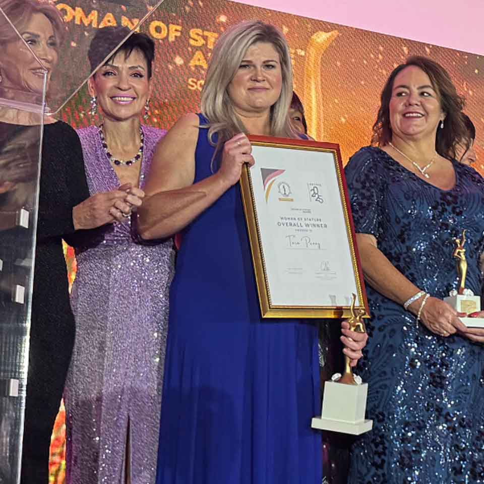 Four women in formal dresses stand on stage at an awards event. The woman in the center, wearing a blue dress, holds a large framed certificate, while another woman beside her holds a trophy. All are smiling.