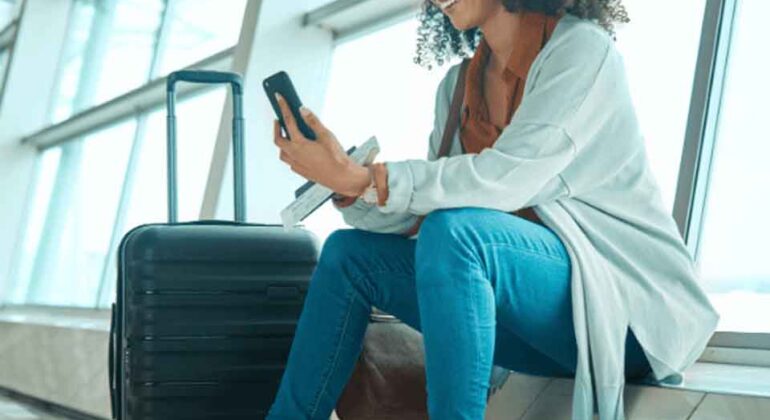 A woman sits in an airport terminal next to a suitcase, smiling while looking at her phone. She is casually dressed in jeans, boots, and a cardigan, with large windows and natural light in the background.