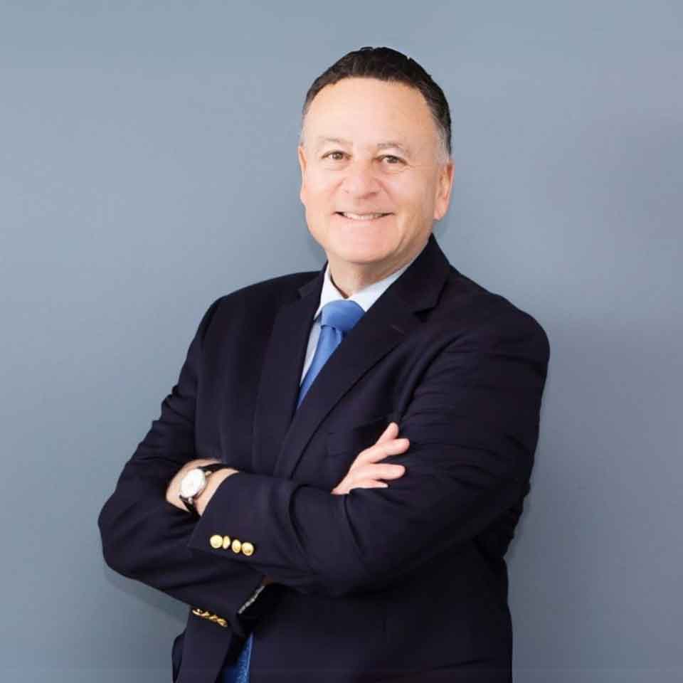 A man in a dark suit, blue tie, and white shirt stands with arms crossed, smiling in front of a plain gray background.