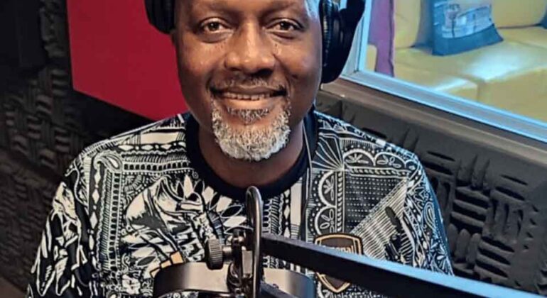 A smiling man with headphones and a patterned shirt sits in a recording studio in front of a microphone, with soundproof foam on the walls and a window showing a yellow sofa in the background.