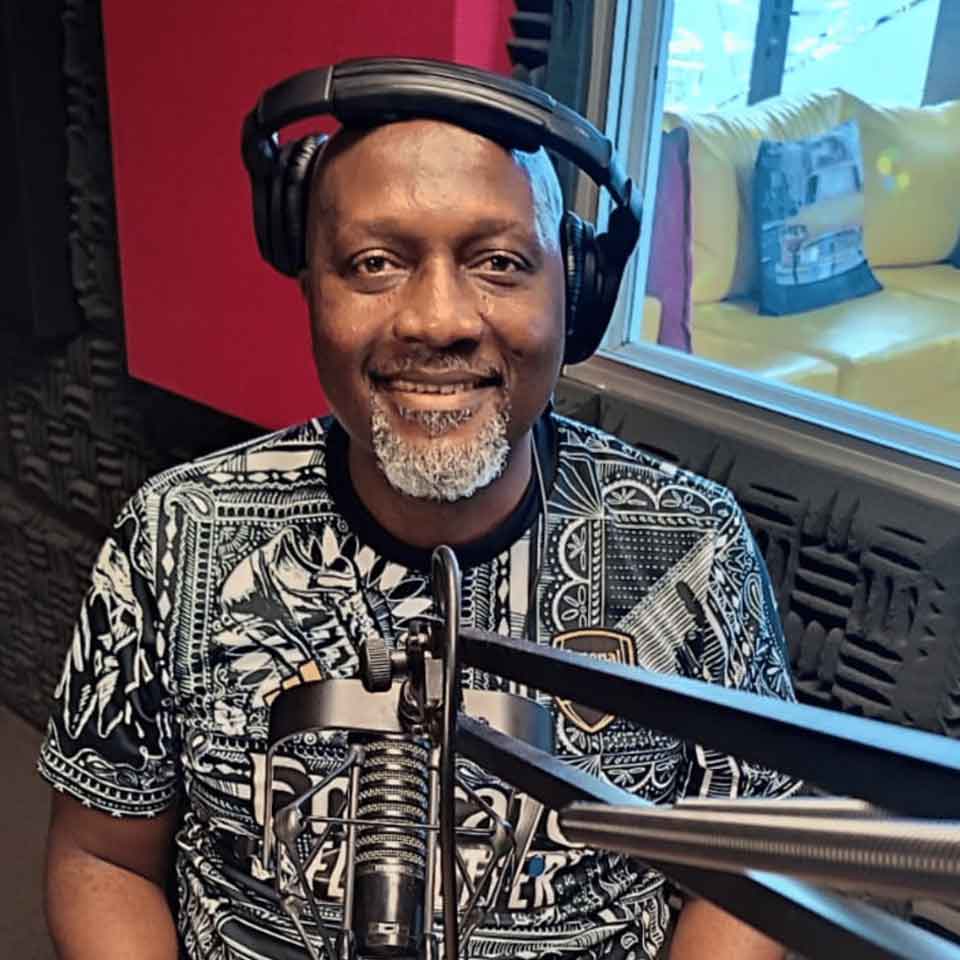 A smiling man with headphones and a patterned shirt sits in a recording studio in front of a microphone, with soundproof foam on the walls and a window showing a yellow sofa in the background.