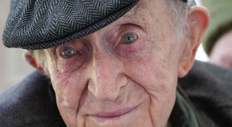 Elderly man wearing a patterned flat cap and dark jacket, smiling gently at the camera. His blue eyes and expressive face show signs of age and wisdom.