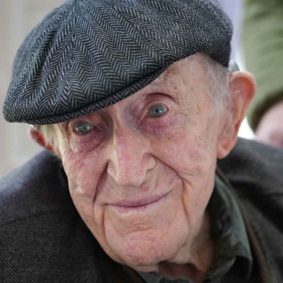 Elderly man wearing a patterned flat cap and dark jacket, smiling gently at the camera. His blue eyes and expressive face show signs of age and wisdom.
