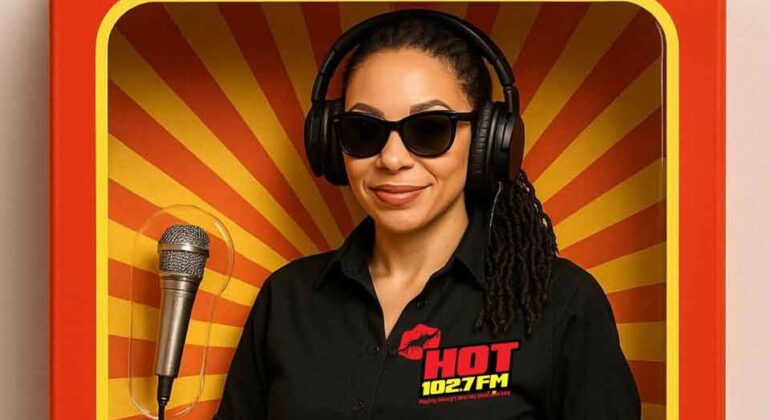 A woman wearing sunglasses, headphones, and a black HOT 102.7 FM shirt stands holding microphones inside a display box with a red and yellow sunburst background, resembling an action figure package.