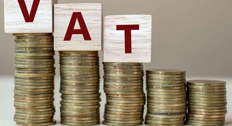 Stacks of coins increase in height from right to left, with wooden blocks on top spelling VAT in red letters, symbolizing value-added tax or financial growth.