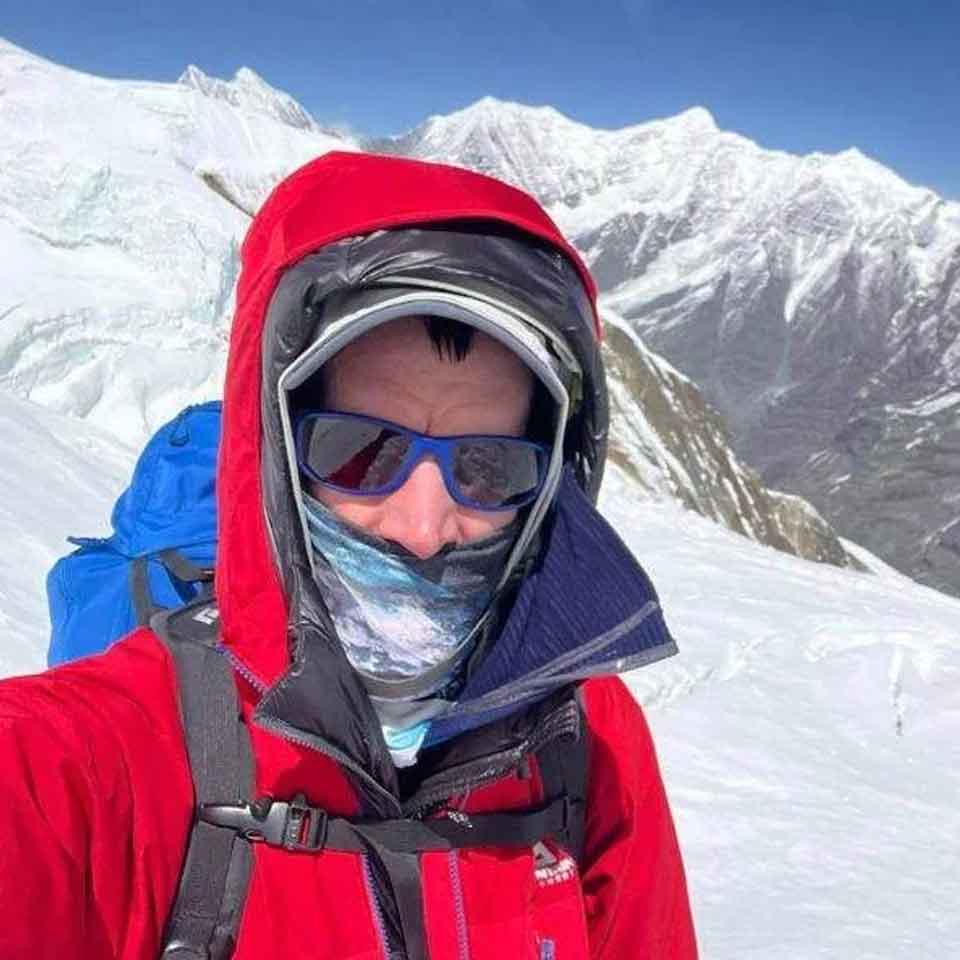A climber wearing a red jacket, blue backpack, sunglasses, and face covering stands on a snow-covered mountain with high peaks and a clear blue sky in the background.