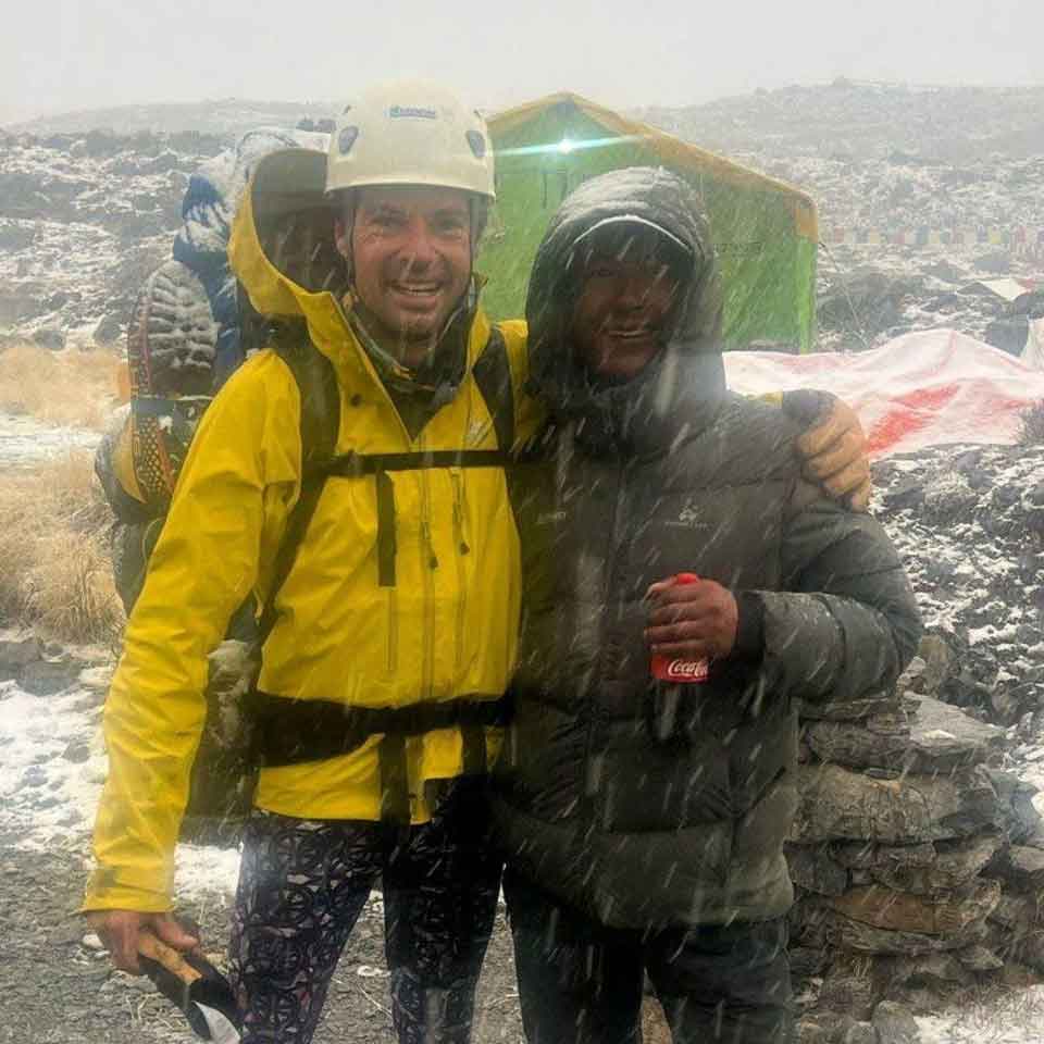 Two people in winter gear stand close together in snowy, windy conditions outdoors. One person wears a yellow jacket, climbing helmet, and backpack; the other wears a dark jacket and holds a Coca-Cola can. A tent is visible in the background.