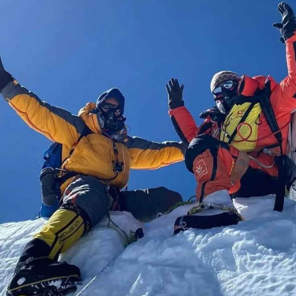 Two climbers in heavy winter gear and oxygen masks sit atop a snowy peak, raising their arms in celebration under a clear blue sky.