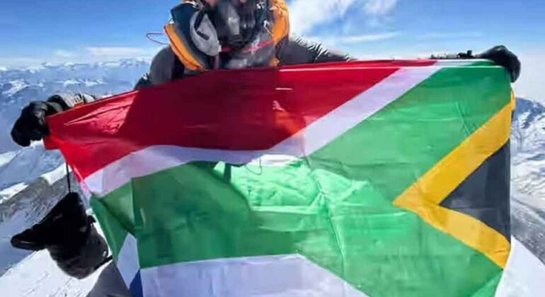 A person wearing cold-weather gear and an oxygen mask holds the South African flag atop a snowy mountain peak, with a clear blue sky and distant mountains in the background.