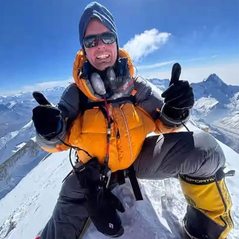 A climber wearing a yellow jacket, sunglasses, and gloves gives two thumbs up while kneeling on the snowy Annapurna Mountain peak with clear blue skies and distant mountain ranges in the background.
