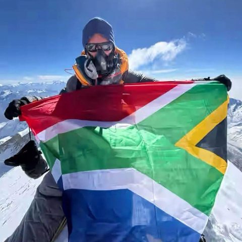 A climber wearing cold weather gear and an oxygen mask holds up the South African flag at the snowy summit of Annapurna Mountain, with a clear blue sky and surrounding snowy peaks in the background.