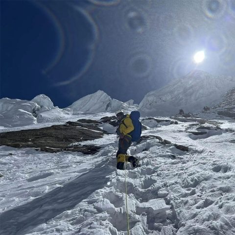 A climber in yellow gear ascends a snowy, steep Annapurna Mountain slope under bright sunlight and a clear blue sky, using a rope for support. Snow-covered peaks and rocky terrain are visible in the background.