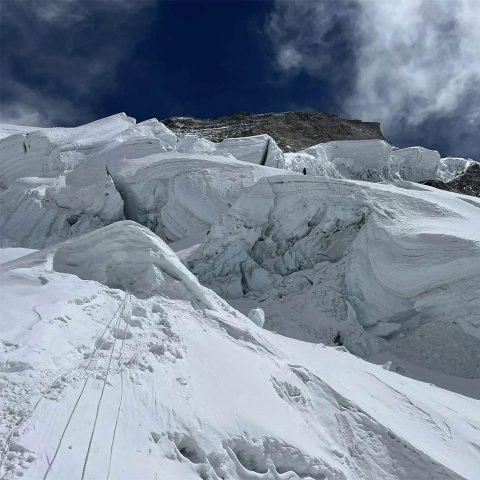 Snowy, jagged ice formations and crevasses line the steep slope of Annapurna Mountain under a dramatic sky, with small climbers visible in the distance and ropes leading up the slope.