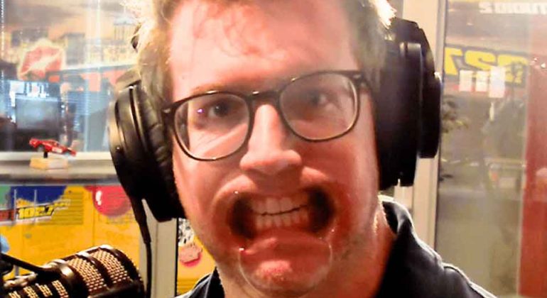 A man with glasses and headphones sits in front of a microphone, pressing his face against a clear surface, distorting his mouth and cheeks in a humorous way.