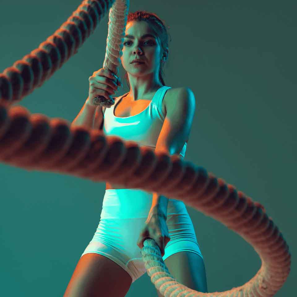 A woman in athletic wear intensely exercises with battle ropes, her face focused and determined, against a teal and orange-lit background.