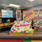 A display of board games on a wooden surface, including 30 Seconds, Jenga, Watch Ya Mouth, Monopoly, Grab & Go, and Twister, with shelves and store merchandise visible in the background.