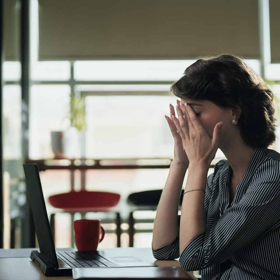 A woman sitting at a desk with a laptop, covering her face with her hands in frustration or exhaustion. A red mug is on the table, and the background shows large windows with natural light.
