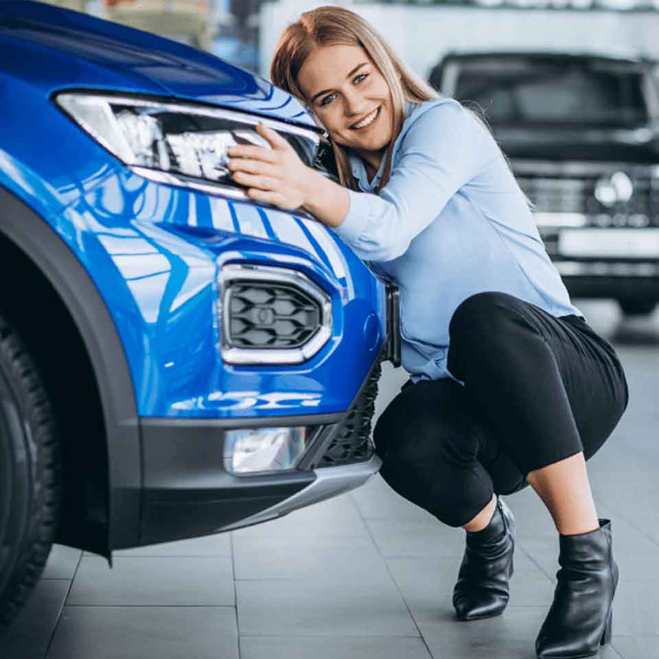 A smiling woman crouches next to a blue car, hugging its front and resting her head near the headlight inside a showroom.