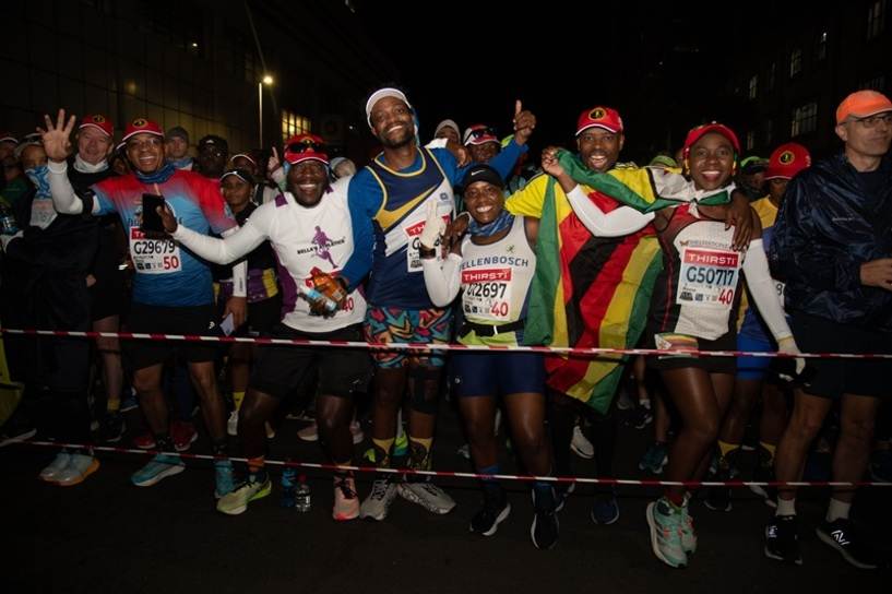 A group of enthusiastic marathon runners, some holding a Zimbabwean flag, smile and pose for the camera at the starting line during a nighttime Comrades Marathon race.