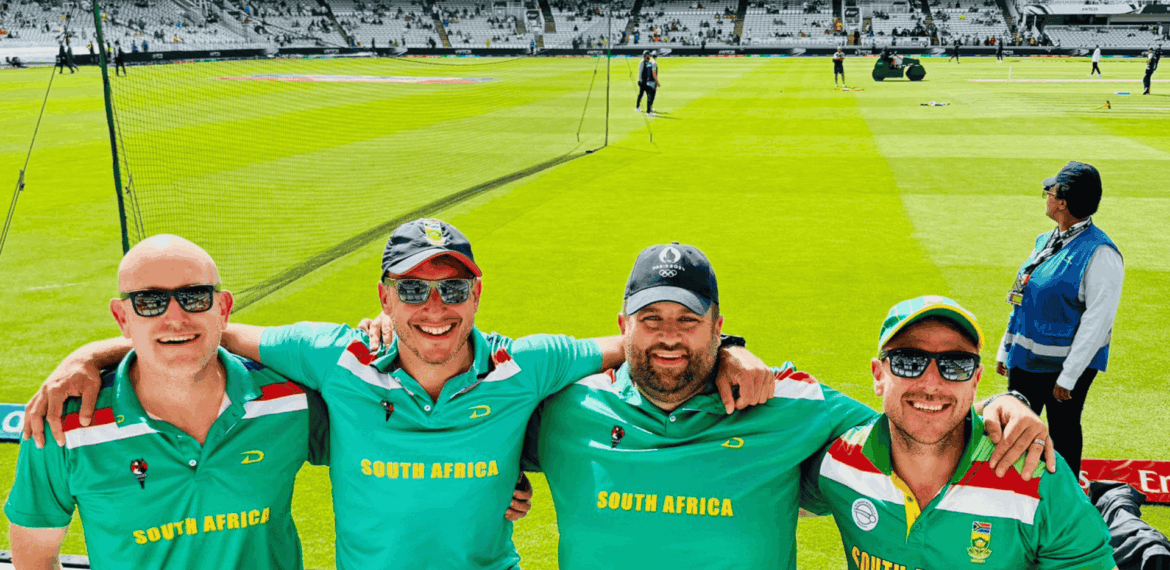 Four smiling men wearing South Africa Proteas cricket shirts pose arm-in-arm at a cricket stadium, with bright green grass, equipment, and other people visible in the background.